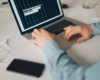 A person is typing on a silver laptop with a screen displaying text about designing digital solutions for big customers. Nearby, there is a black smartphone and white earphones placed on the desk. The person's sleeves are light blue, suggesting a casual attire.