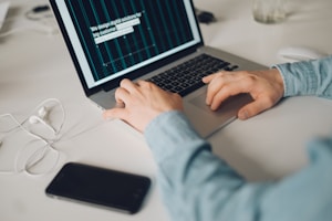 A person is typing on a silver laptop with a screen displaying text about designing digital solutions for big customers. Nearby, there is a black smartphone and white earphones placed on the desk. The person's sleeves are light blue, suggesting a casual attire.
