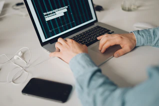 A person is typing on a silver laptop with a screen displaying text about designing digital solutions for big customers. Nearby, there is a black smartphone and white earphones placed on the desk. The person's sleeves are light blue, suggesting a casual attire.