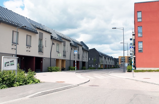 A modern urban setting with rows of townhouses featuring slanted roofs and neutral facades. The street is empty, and the sky is partly cloudy. A large red building stands prominently on the right, contrasting with the muted tones of the townhouses.