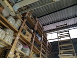 A storage area with wooden shelves filled with large containers, boxes, and miscellaneous items. The shelves are arranged vertically and extend up to the corrugated metal roof. A wooden ladder leans against the shelves, providing access to higher levels. There is natural light coming in through a translucent panel in the roof.