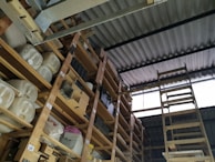 A storage area with wooden shelves filled with large containers, boxes, and miscellaneous items. The shelves are arranged vertically and extend up to the corrugated metal roof. A wooden ladder leans against the shelves, providing access to higher levels. There is natural light coming in through a translucent panel in the roof.