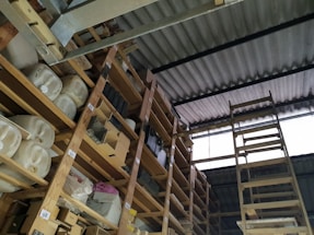 A storage area with wooden shelves filled with large containers, boxes, and miscellaneous items. The shelves are arranged vertically and extend up to the corrugated metal roof. A wooden ladder leans against the shelves, providing access to higher levels. There is natural light coming in through a translucent panel in the roof.