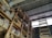 A storage area with wooden shelves filled with large containers, boxes, and miscellaneous items. The shelves are arranged vertically and extend up to the corrugated metal roof. A wooden ladder leans against the shelves, providing access to higher levels. There is natural light coming in through a translucent panel in the roof.