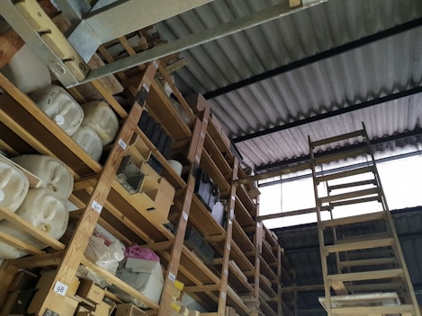 A storage area with wooden shelves filled with large containers, boxes, and miscellaneous items. The shelves are arranged vertically and extend up to the corrugated metal roof. A wooden ladder leans against the shelves, providing access to higher levels. There is natural light coming in through a translucent panel in the roof.