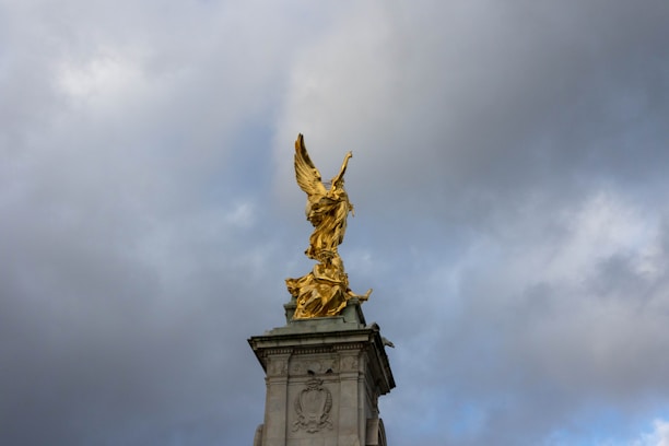 A golden statue of an angel holding a laurel wreath stands atop a stone monument. The sky in the background is overcast with dark clouds, creating a dramatic contrast with the bright gold of the statue.