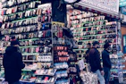 A bustling market stall is filled with a wide array of mobile phone cases and accessories, displayed on shelves and racks. Several people are browsing the items, dressed warmly for the weather. The cases come in various colors and designs, adding vibrancy to the scene.
