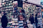 A bustling market stall is filled with a wide array of mobile phone cases and accessories, displayed on shelves and racks. Several people are browsing the items, dressed warmly for the weather. The cases come in various colors and designs, adding vibrancy to the scene.