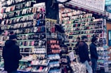 A bustling market stall is filled with a wide array of mobile phone cases and accessories, displayed on shelves and racks. Several people are browsing the items, dressed warmly for the weather. The cases come in various colors and designs, adding vibrancy to the scene.