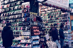 A bustling market stall is filled with a wide array of mobile phone cases and accessories, displayed on shelves and racks. Several people are browsing the items, dressed warmly for the weather. The cases come in various colors and designs, adding vibrancy to the scene.