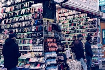 A bustling market stall is filled with a wide array of mobile phone cases and accessories, displayed on shelves and racks. Several people are browsing the items, dressed warmly for the weather. The cases come in various colors and designs, adding vibrancy to the scene.