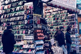 A bustling market stall is filled with a wide array of mobile phone cases and accessories, displayed on shelves and racks. Several people are browsing the items, dressed warmly for the weather. The cases come in various colors and designs, adding vibrancy to the scene.