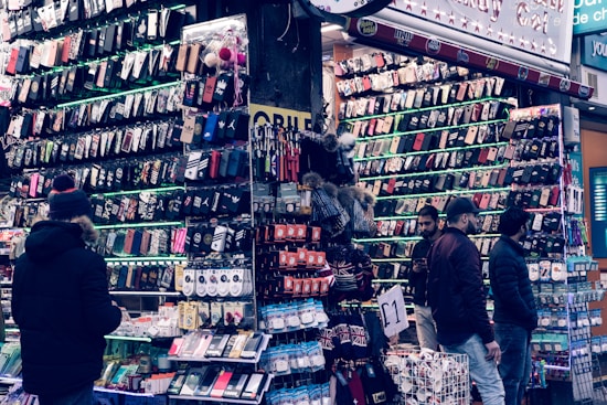 A bustling market stall is filled with a wide array of mobile phone cases and accessories, displayed on shelves and racks. Several people are browsing the items, dressed warmly for the weather. The cases come in various colors and designs, adding vibrancy to the scene.
