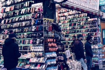 A bustling market stall is filled with a wide array of mobile phone cases and accessories, displayed on shelves and racks. Several people are browsing the items, dressed warmly for the weather. The cases come in various colors and designs, adding vibrancy to the scene.