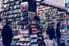 A bustling market stall is filled with a wide array of mobile phone cases and accessories, displayed on shelves and racks. Several people are browsing the items, dressed warmly for the weather. The cases come in various colors and designs, adding vibrancy to the scene.