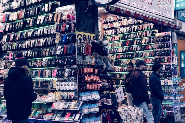 A bustling market stall is filled with a wide array of mobile phone cases and accessories, displayed on shelves and racks. Several people are browsing the items, dressed warmly for the weather. The cases come in various colors and designs, adding vibrancy to the scene.