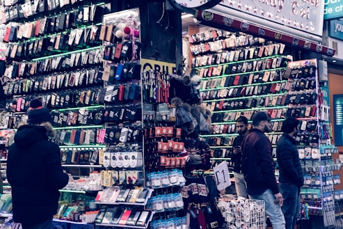 A bustling market stall is filled with a wide array of mobile phone cases and accessories, displayed on shelves and racks. Several people are browsing the items, dressed warmly for the weather. The cases come in various colors and designs, adding vibrancy to the scene.