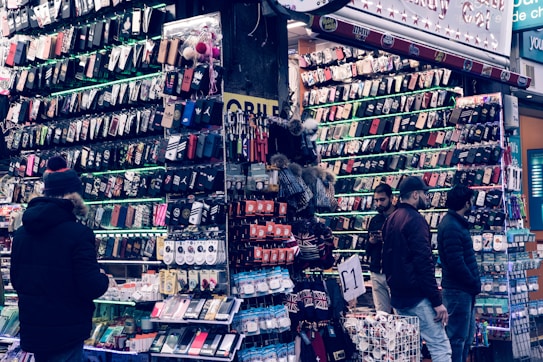 A bustling market stall is filled with a wide array of mobile phone cases and accessories, displayed on shelves and racks. Several people are browsing the items, dressed warmly for the weather. The cases come in various colors and designs, adding vibrancy to the scene.
