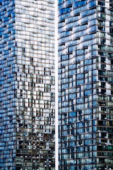 Two high-rise buildings with a modern, glass facade reflecting light, creating a mosaic of blue and gray tones. The architectural design features a wavy pattern with varying depths and textures, contributing to a dynamic visual effect.