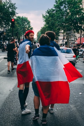 A group of people walking down a street, one of whom is wearing a red beret. Two individuals are wrapped in a large flag colored blue, white, and red. Cars and other pedestrians are visible in the background, along with trees lining the street.
