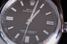 Close-up view of a luxury wristwatch featuring a sleek black dial with silver hour markers and hands, encased in a polished stainless steel. The branding and model information are visible on the dial, emphasizing elegance and precision.