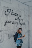 A person with a beard and wearing a denim jacket and a headband is reading a magazine in front of a large mural. The mural features line drawings of people engaged in various activities and a prominent phrase 'Home is where your story begins.' The background includes drawings of mountains and a yurt.