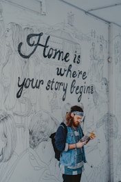 A person with a beard and wearing a denim jacket and a headband is reading a magazine in front of a large mural. The mural features line drawings of people engaged in various activities and a prominent phrase 'Home is where your story begins.' The background includes drawings of mountains and a yurt.