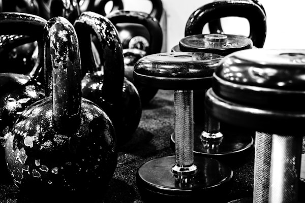 A set of heavy-duty dumbbells and kettlebells lined up on a gym floor with strong lighting.