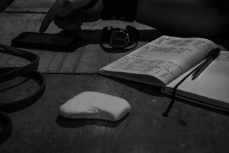 A monochrome scene with a notebook open to handwritten notes, a pen placed on top, and a smartphone nearby. A pointing hand is visible near the smartphone. There is also a watch and an unknown white object on the table, possibly a piece of paper.