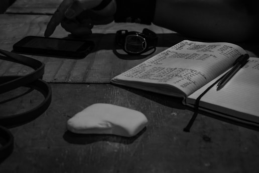 A monochrome scene with a notebook open to handwritten notes, a pen placed on top, and a smartphone nearby. A pointing hand is visible near the smartphone. There is also a watch and an unknown white object on the table, possibly a piece of paper.