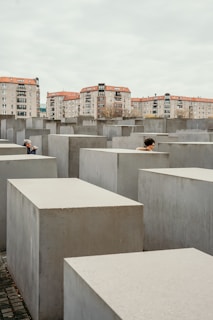 A series of gray concrete slabs arranged in a grid pattern, with two people partially visible among the slabs. In the background, there are several residential buildings with red roofs under a cloudy sky.