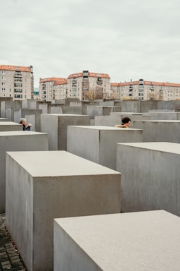 A series of gray concrete slabs arranged in a grid pattern, with two people partially visible among the slabs. In the background, there are several residential buildings with red roofs under a cloudy sky.