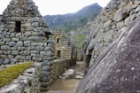Stone structures featuring ancient, Incan architectural design with terraced hillsides in the background. The setting is mountainous and lush with greenery.