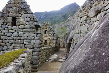 Stone structures featuring ancient, Incan architectural design with terraced hillsides in the background. The setting is mountainous and lush with greenery.