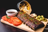 A wooden platter features a rack of glazed ribs garnished with herbs. Sliced tomatoes, a small bowl of barbecue sauce, and pickled cucumber slices are arranged around the ribs. Crispy fries spill out of a metallic bucket in the background, accompanied by a baked potato with cheese and chives.