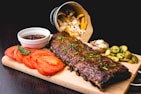 A wooden platter features a rack of glazed ribs garnished with herbs. Sliced tomatoes, a small bowl of barbecue sauce, and pickled cucumber slices are arranged around the ribs. Crispy fries spill out of a metallic bucket in the background, accompanied by a baked potato with cheese and chives.