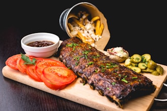 A wooden platter features a rack of glazed ribs garnished with herbs. Sliced tomatoes, a small bowl of barbecue sauce, and pickled cucumber slices are arranged around the ribs. Crispy fries spill out of a metallic bucket in the background, accompanied by a baked potato with cheese and chives.