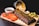 A wooden platter features a rack of glazed ribs garnished with herbs. Sliced tomatoes, a small bowl of barbecue sauce, and pickled cucumber slices are arranged around the ribs. Crispy fries spill out of a metallic bucket in the background, accompanied by a baked potato with cheese and chives.