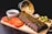 A wooden platter features a rack of glazed ribs garnished with herbs. Sliced tomatoes, a small bowl of barbecue sauce, and pickled cucumber slices are arranged around the ribs. Crispy fries spill out of a metallic bucket in the background, accompanied by a baked potato with cheese and chives.