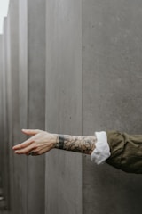 An arm with tattoos, wearing a dark green sleeve, extends outward against a backdrop of large, smooth gray concrete blocks. The tattoos depict floral designs, and the hand is slightly open.