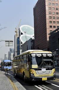 A busy urban street features a large yellow airport limousine bus in the foreground, with a distinctive modern high-rise building featuring unusual geometric shapes under construction in the background. A blue city bus and other high-rise buildings are visible alongside a yellow-striped road, creating a dynamic and bustling atmosphere.