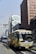 A busy urban street features a large yellow airport limousine bus in the foreground, with a distinctive modern high-rise building featuring unusual geometric shapes under construction in the background. A blue city bus and other high-rise buildings are visible alongside a yellow-striped road, creating a dynamic and bustling atmosphere.