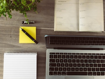 A wooden desk displays an open planner, a laptop, a notepad, a yellow sticky note with a pen, and a small potted plant. The planner is dated December 2016 and lies open to a week view. The laptop is positioned towards the bottom of the image with its keyboard visible.