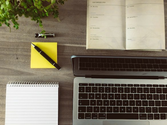 A wooden desk displays an open planner, a laptop, a notepad, a yellow sticky note with a pen, and a small potted plant. The planner is dated December 2016 and lies open to a week view. The laptop is positioned towards the bottom of the image with its keyboard visible.