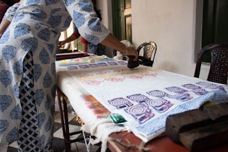A person is engaged in block printing on fabric spread across a table. The fabric displays colorful, intricate patterns created with wooden blocks. The person is wearing a white and blue printed garment, matching the motif being applied on the fabric.