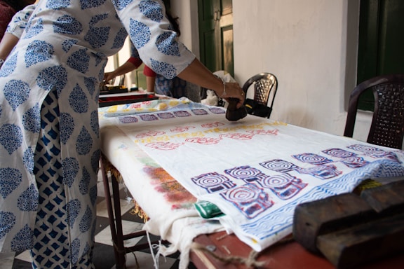 A person is engaged in block printing on fabric spread across a table. The fabric displays colorful, intricate patterns created with wooden blocks. The person is wearing a white and blue printed garment, matching the motif being applied on the fabric.