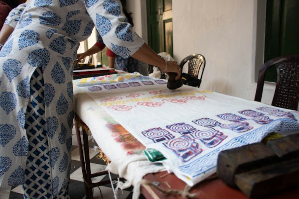 A person is engaged in block printing on fabric spread across a table. The fabric displays colorful, intricate patterns created with wooden blocks. The person is wearing a white and blue printed garment, matching the motif being applied on the fabric.