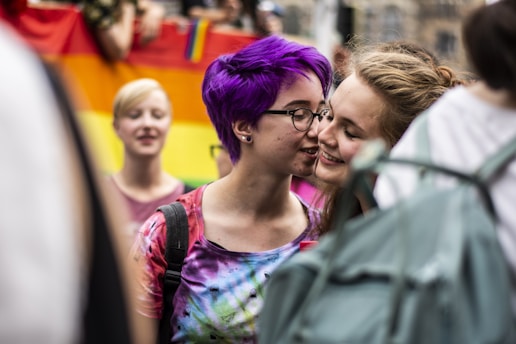 Two individuals share a close, intimate moment in a colorful, public setting. One person has vibrant purple hair and wears glasses, while the other is smiling warmly. In the background, a rainbow flag suggests a celebration of diversity and inclusivity.