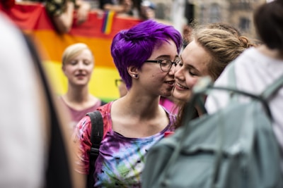 Two individuals share a close, intimate moment in a colorful, public setting. One person has vibrant purple hair and wears glasses, while the other is smiling warmly. In the background, a rainbow flag suggests a celebration of diversity and inclusivity.