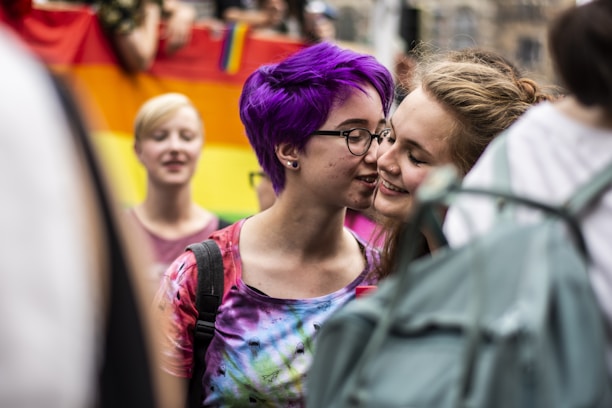 Two individuals share a close, intimate moment in a colorful, public setting. One person has vibrant purple hair and wears glasses, while the other is smiling warmly. In the background, a rainbow flag suggests a celebration of diversity and inclusivity.